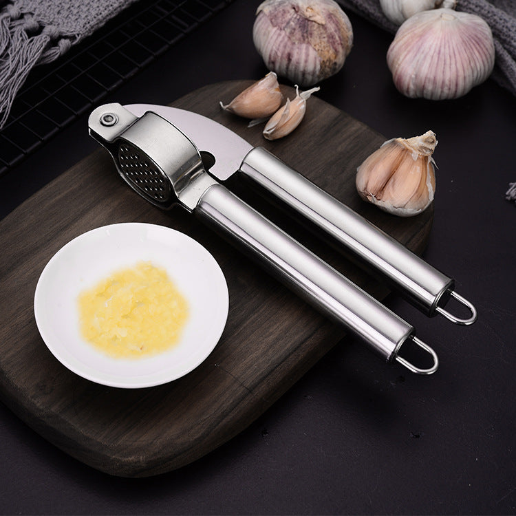 Stainless steel garlic press on a wooden cutting board beside minced garlic and bulbs in a kitchen; props not included.
