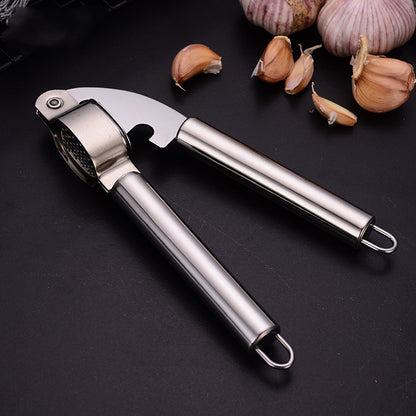 Stainless steel garlic press resting on a dark surface next to garlic bulbs and peeled cloves; garlic not included.